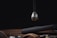 A spherical, futuristic device with a glossy black and silver finish, partially suspended. It has yellow accents and numbers, resembling a timer or measuring tool. Beneath it, a wooden surface holds a brown leather strap and dark metallic objects, suggesting a high-tech or mechanical setting.