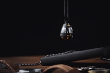 A spherical, futuristic device with a glossy black and silver finish, partially suspended. It has yellow accents and numbers, resembling a timer or measuring tool. Beneath it, a wooden surface holds a brown leather strap and dark metallic objects, suggesting a high-tech or mechanical setting.