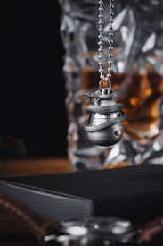 A close-up view of a silver keychain pendant shaped like a grenade, wrapped with a textured snake design, hanging on a ball chain. The background features a glass with an amber liquid, possibly whiskey, and a black leather-bound notebook near a wristwatch with a brown leather strap.
