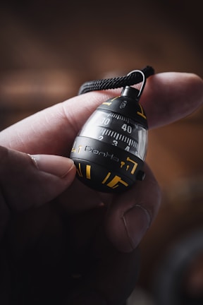 A close-up of a hand holding a small, round device with a black and yellow design. The device features numbers, possibly indicating some form of measurement or countdown, and a strap attached to its top.