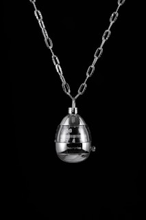 A shiny metal egg-shaped timer hangs from a chain against a black background. The timer has clearly marked numbers and a mechanical knob for setting time.