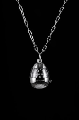 A shiny metal egg-shaped timer hangs from a chain against a black background. The timer has clearly marked numbers and a mechanical knob for setting time.