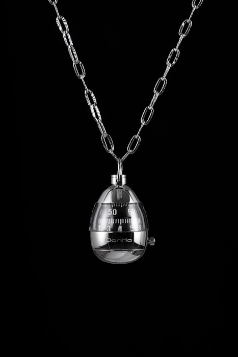 A shiny metal egg-shaped timer hangs from a chain against a black background. The timer has clearly marked numbers and a mechanical knob for setting time.