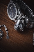 A high-quality wristwatch with a metallic chain bracelet is paired with an intricate, modern-looking keychain that resembles a mechanical object. These items are set against a dark wooden surface, providing a rich contrast to the polished metal of the watch and keychain.