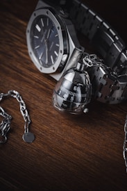 A high-quality wristwatch with a metallic chain bracelet is paired with an intricate, modern-looking keychain that resembles a mechanical object. These items are set against a dark wooden surface, providing a rich contrast to the polished metal of the watch and keychain.