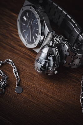 A high-quality wristwatch with a metallic chain bracelet is paired with an intricate, modern-looking keychain that resembles a mechanical object. These items are set against a dark wooden surface, providing a rich contrast to the polished metal of the watch and keychain.