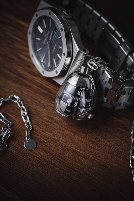 A high-quality wristwatch with a metallic chain bracelet is paired with an intricate, modern-looking keychain that resembles a mechanical object. These items are set against a dark wooden surface, providing a rich contrast to the polished metal of the watch and keychain.