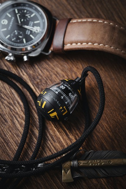 A sophisticated flat lay of a leather-strapped wristwatch, featuring a detailed black and silver dial. Beside it sits a metallic gyroscope or spinning top-like object adorned with yellow markings, entwined by a black cord. There's also a decorative feather and stylized dagger motif. The composition is set on a textured wooden surface, enhancing the classy and rugged aesthetic.