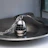 A metallic egg-shaped kitchen timer with a sleek design sits on a silver dish. The timer features a black rotating dial with white numbers and a decorative chain attached to the top. The image has a reflective surface and is artistically composed with soft light.