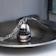 A metallic egg-shaped kitchen timer with a sleek design sits on a silver dish. The timer features a black rotating dial with white numbers and a decorative chain attached to the top. The image has a reflective surface and is artistically composed with soft light.