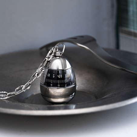 A metallic egg-shaped kitchen timer with a sleek design sits on a silver dish. The timer features a black rotating dial with white numbers and a decorative chain attached to the top. The image has a reflective surface and is artistically composed with soft light.