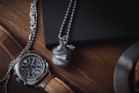 A luxurious wristwatch with a brown leather strap and a detailed face is placed on a wooden surface. Next to it, a metallic ball pendant with a snake design wraps around it. Both items are situated near a dark-colored notebook and part of a metal object, possibly a cup or plate.