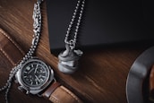 A luxurious wristwatch with a brown leather strap and a detailed face is placed on a wooden surface. Next to it, a metallic ball pendant with a snake design wraps around it. Both items are situated near a dark-colored notebook and part of a metal object, possibly a cup or plate.