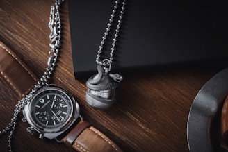 A luxurious wristwatch with a brown leather strap and a detailed face is placed on a wooden surface. Next to it, a metallic ball pendant with a snake design wraps around it. Both items are situated near a dark-colored notebook and part of a metal object, possibly a cup or plate.