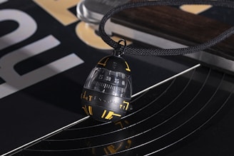 A close-up view of a black and gold pendant hanging from a black cord, resting on a turntable. The pendant has numbers and symbols in yellow and a sleek, modern design. There are abstract reflections on the shiny surface of the turntable, and portions of a printed design are visible in the background.