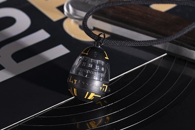 A close-up view of a black and gold pendant hanging from a black cord, resting on a turntable. The pendant has numbers and symbols in yellow and a sleek, modern design. There are abstract reflections on the shiny surface of the turntable, and portions of a printed design are visible in the background.