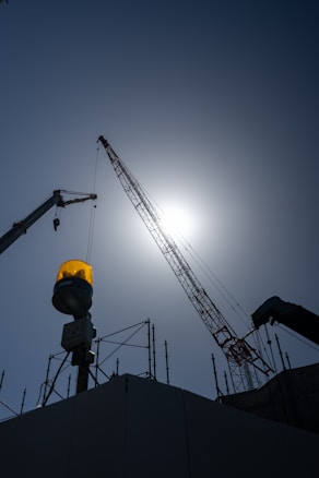 A construction site with a prominent crane extending into the sky. The sun is directly behind the crane, creating a silhouette effect. An amber warning light is visible, and scaffolding surrounds the area.