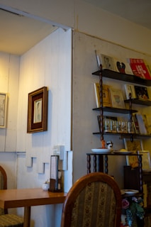 A cozy interior space featuring a small wooden table with a patterned chair. On the wall, there are several shelves holding books, framed art, and decorative items. The lighting is warm, contributing to a comfortable atmosphere.