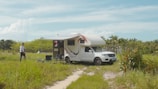 A white camper van is parked in a grassy field with an awning extended and outdoor chairs set up beside it. A person is walking towards the van from the left. The surroundings are filled with lush green vegetation and palm trees, under a clear blue sky with a few clouds.