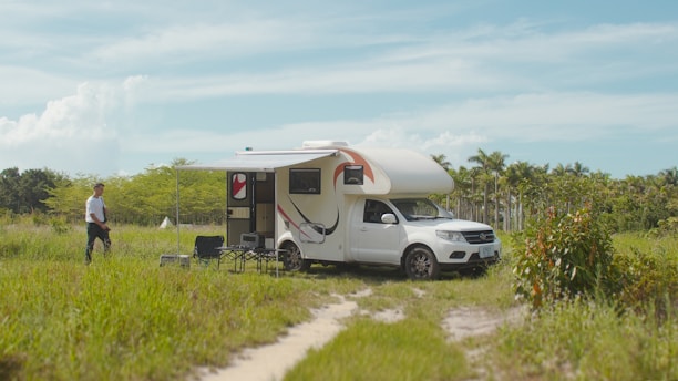 A white camper van is parked in a grassy field with an awning extended and outdoor chairs set up beside it. A person is walking towards the van from the left. The surroundings are filled with lush green vegetation and palm trees, under a clear blue sky with a few clouds.