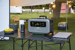 A portable power station is placed on a black, foldable table outdoors. The device has a digital display and appears to be modern and robust. Surrounding objects include a bowl of fruits, a portable stove, a tumbler, a laptop, and a smartphone. There is a camping setup with string lights, a tent, and some chairs in the background, on a grassy area with trees.