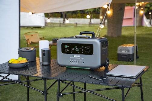 A portable power station is placed on a black, foldable table outdoors. The device has a digital display and appears to be modern and robust. Surrounding objects include a bowl of fruits, a portable stove, a tumbler, a laptop, and a smartphone. There is a camping setup with string lights, a tent, and some chairs in the background, on a grassy area with trees.