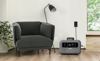 A modern living space featuring a dark green armchair next to a tall black standing lamp. A gray portable power station is placed on the hardwood floor, plugged into a wall socket. Adjacent to the armchair is a leafy green plant, and a window reveals a glimpse of greenery outside.