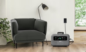 A modern living space featuring a dark green armchair next to a tall black standing lamp. A gray portable power station is placed on the hardwood floor, plugged into a wall socket. Adjacent to the armchair is a leafy green plant, and a window reveals a glimpse of greenery outside.