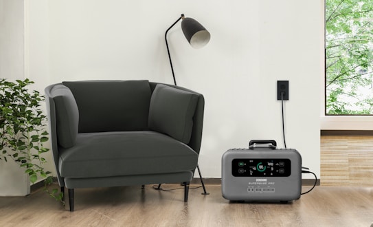 A modern living space featuring a dark green armchair next to a tall black standing lamp. A gray portable power station is placed on the hardwood floor, plugged into a wall socket. Adjacent to the armchair is a leafy green plant, and a window reveals a glimpse of greenery outside.