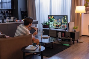 A living room setup with a person sitting on a couch, holding a remote, facing a television screen displaying a video game. The room features a modern design with a kitchen area visible in the background, including a countertop with fruits and appliances. The television stands on a media console beside a potted plant and a lamp, creating a cozy and relaxed atmosphere.