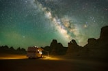 A motorhome is parked in a desert landscape under a sky filled with the Milky Way galaxy. The motorhome is illuminated, contrasting with the dark silhouettes of rocky formations around it.