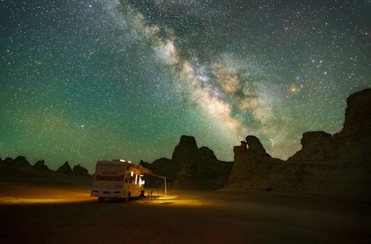 A motorhome is parked in a desert landscape under a sky filled with the Milky Way galaxy. The motorhome is illuminated, contrasting with the dark silhouettes of rocky formations around it.