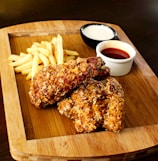 A wooden platter holds two pieces of crispy fried chicken next to a small serving of golden French fries. Accompanying the meal are two dips in separate small bowls, one appears to be a creamy white sauce while the other is a reddish sauce, possibly ketchup or hot sauce.