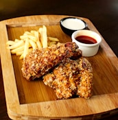 A wooden platter holds two pieces of crispy fried chicken next to a small serving of golden French fries. Accompanying the meal are two dips in separate small bowls, one appears to be a creamy white sauce while the other is a reddish sauce, possibly ketchup or hot sauce.