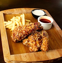 A wooden platter holds two pieces of crispy fried chicken next to a small serving of golden French fries. Accompanying the meal are two dips in separate small bowls, one appears to be a creamy white sauce while the other is a reddish sauce, possibly ketchup or hot sauce.