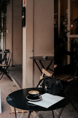 A small, round black table at an outdoor cafe with a cup of coffee featuring latte art, a menu, and a black bag placed on it. Wooden chairs with metal frames are positioned around the table, and the setting suggests a minimalist and cozy ambiance. The background shows part of a cafe entrance with a glass door.