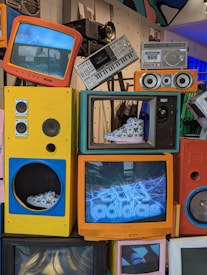 A colorful assortment of retro electronics, including televisions, speakers, and a keyboard, displayed in an artistic manner. Some of the TVs showcase sneakers inside their frames. The background includes additional electronic devices and parts, creating a nostalgic yet vibrant atmosphere.