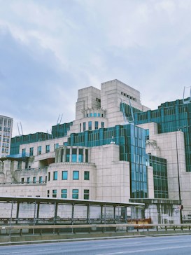 A large, modern building with distinct, geometric architecture featuring a combination of cream-colored stone and teal-tinted glass. The sky is overcast, creating a subdued atmosphere. The structure includes multiple levels and angular projections, with some parts resembling a fortress.