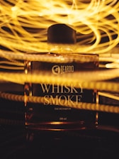 A perfume bottle labeled 'WHISKY SMOKE' is illuminated with warm, swirling golden lights in the background, creating an artistic and vibrant effect.