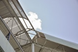 A metallic structure with a slatted roof and supporting beams, seen from an angle against a clear blue sky with a small cloud.