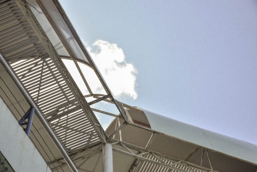 A metallic structure with a slatted roof and supporting beams, seen from an angle against a clear blue sky with a small cloud.