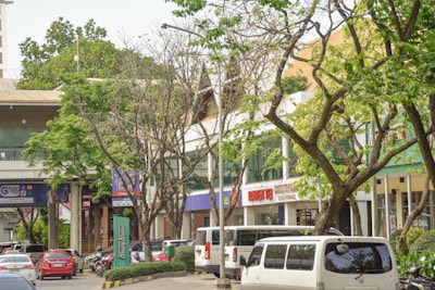 A bustling urban environment with a mix of modern buildings and lush trees lining the street. Vehicles, including cars and vans, are parked and moving along the road. Various businesses with signage are visible, contributing to a lively city atmosphere.