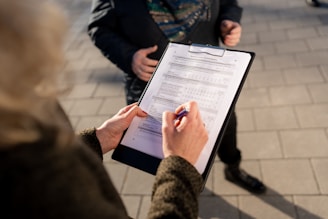 A person is holding a clipboard with a form attached and filling it out with a pen. Another person's torso is partially visible in the background, suggesting a survey or registration process happening outdoors. The ground is paved with rectangular tiles.