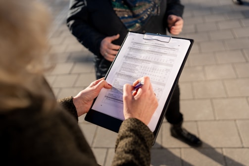 A person is holding a clipboard with a form attached and filling it out with a pen. Another person's torso is partially visible in the background, suggesting a survey or registration process happening outdoors. The ground is paved with rectangular tiles.
