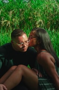 A couple sits closely together in a lush green natural setting. One person is wearing glasses and appears contemplative as the other person leans in to give a gentle kiss on the cheek, conveying affection and warmth.