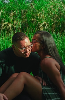 A couple sits closely together in a lush green natural setting. One person is wearing glasses and appears contemplative as the other person leans in to give a gentle kiss on the cheek, conveying affection and warmth.
