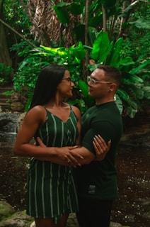 A couple is standing closely together in an intimate embrace in a lush, green forest environment, with a small body of water and dense foliage in the background. Both people are wearing green clothing, and the sunlight softly illuminates their faces, creating an intimate and natural atmosphere.