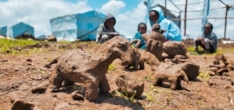 A close-up view of several handmade clay sculptures resembling animals placed on the ground, with a blurred background showing four children in hoodies sitting on grass near blue tents. The setting appears informal and outdoor, suggesting a community or camp environment.