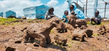 A close-up view of several handmade clay sculptures resembling animals placed on the ground, with a blurred background showing four children in hoodies sitting on grass near blue tents. The setting appears informal and outdoor, suggesting a community or camp environment.