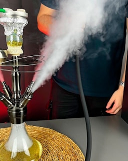 A hookah is emitting smoke as it sits on a round woven mat. The hookah has multiple hoses, and smoke is visibly coming out of several ports. A person nearby is partially visible holding a hose. The atmosphere suggests the hookah is in use, with the smoke clouds creating a soft and slightly dramatic scene.